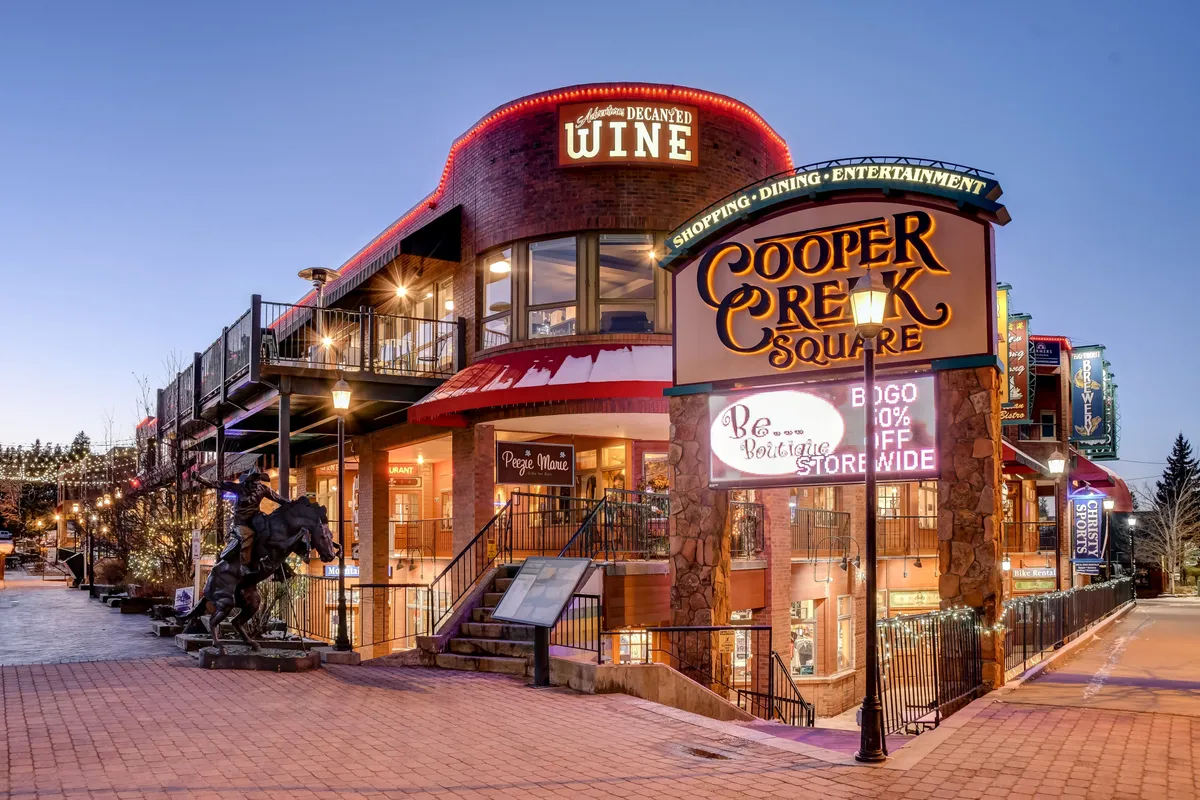 A vibrant exterior shot featuring the Cooper Creek Square sign, charming wine shop signage, and warm exterior lighting. The scene hints at bustling shopping, dining, and entertainment options with an inviting, lively mood.