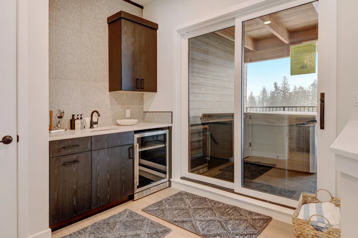A stylish, modern wet bar area indoors with sleek cabinetry and a large sliding glass door leading out to a deck. The space is designed for socializing, with bar seating and ample light streaming in, hinting at seamless indoor-outdoor living.