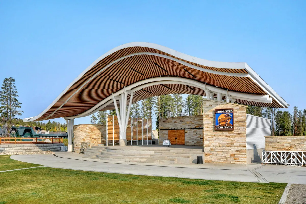 A festive, open-air stage area framed by a striking curved roof structure and a stone facade. The scene is lively, suggesting outdoor concerts and community gatherings, with plenty of space for families to enjoy local events.