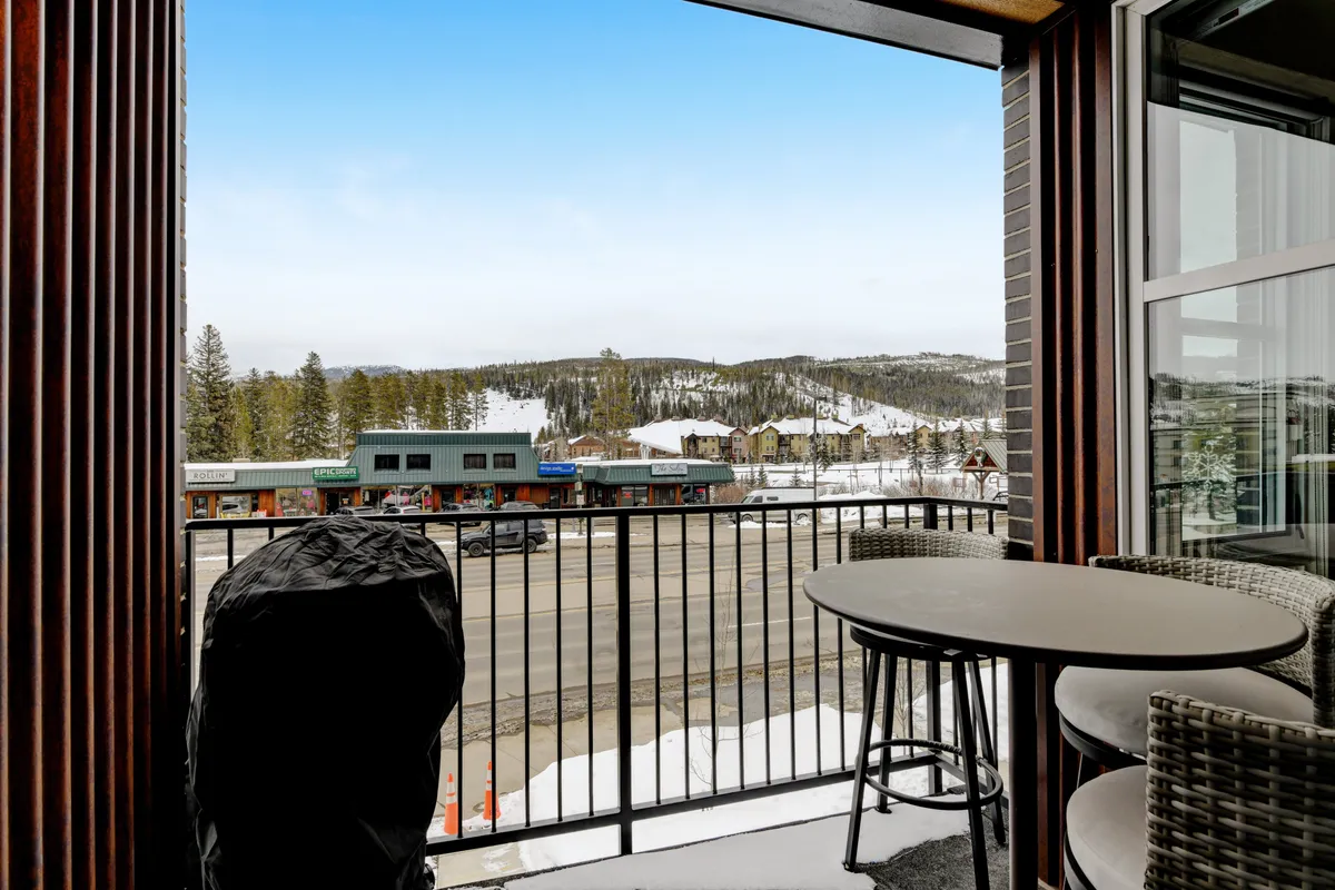 A cheerful balcony scene overlooking snow-covered ground, with comfortable seating, surrounding mountains, and nearby shops visible. People enjoy coffee and socialize as the sun sets, capturing the vibrant, communal energy of the deck while showcasing spectacular Winter Park views.