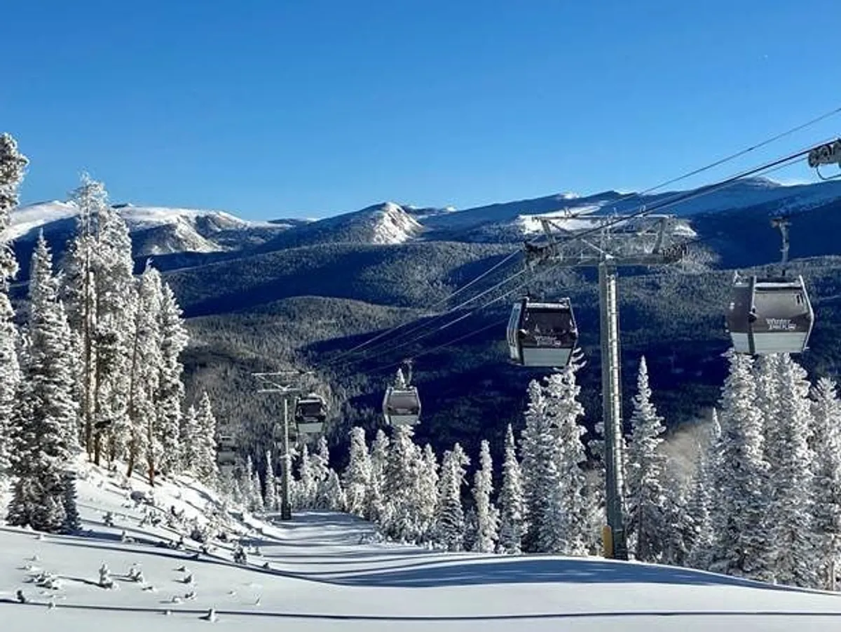 A breathtaking view of a ski lift gliding above snow-laden trees with a dramatic mountain range in the background. The scene evokes a sense of adventure and serenity, capturing the essence of winter recreation such as skiing, snowboarding, and mountain exploration.