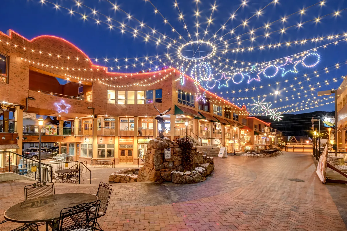 An inviting outdoor plaza with decorative string lights, comfortable seating areas, and a distinctive stone fountain. People are implied to be enjoying outdoor dining and mingling, with a festive and welcoming ambiance.