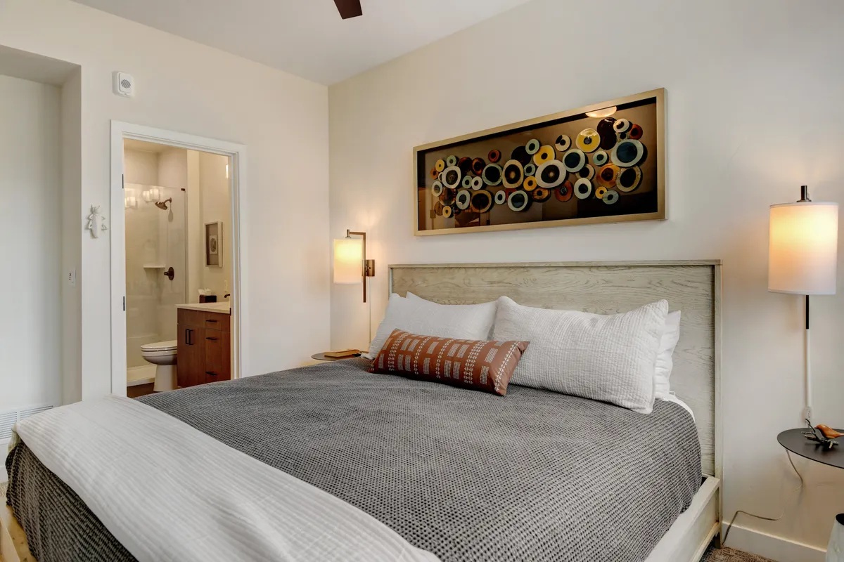 A tranquil bedroom with a king-sized bed topped with decorative pillows, a soft throw, and an art piece above the headboard. Natural light flows through a window, and a nightstand with a lamp sits nearby. A bathroom door is visible in the background, hinting at ensuite convenience.