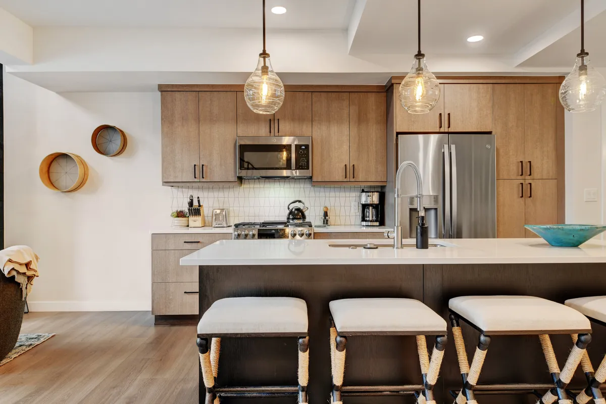 A contemporary kitchen scene with an island countertop, sleek pendant lights, stools for casual breakfasts, ample cabinetry, and high-end appliances. The space is bright and welcoming, designed for both efficiency and gathering.