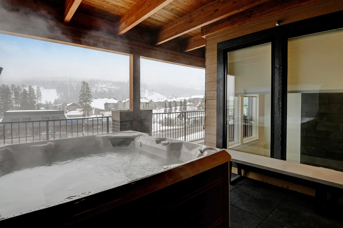 A steaming hot tub nestled on an outdoor deck, surrounded by snow-laden landscape and sturdy wooden beams. The scene captures a tranquil winter environment, with fresh powder in the background and the inviting water promising relaxation after a day of adventure.